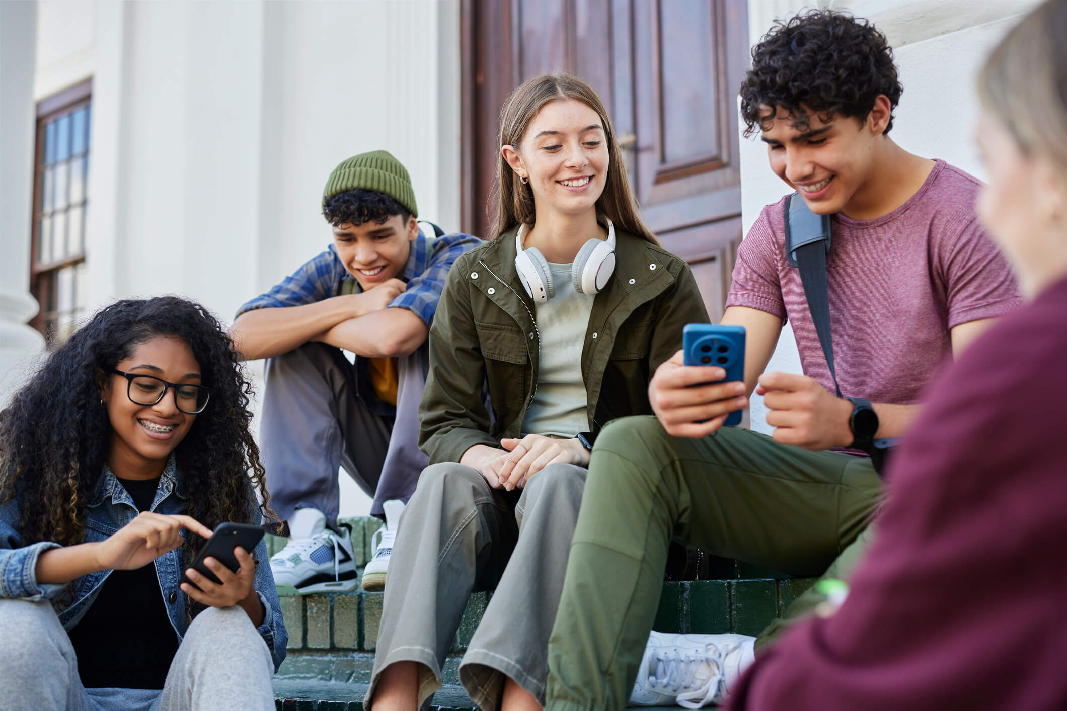 Diverse group of students sitting on outdoor steps, smiling and using smartphones together between classes.